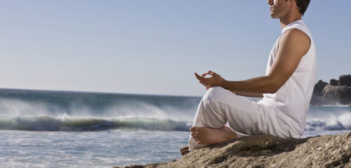 Handsome young man meditating on rock by sea